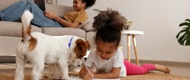 A girl lays on the floor while drawing as her puppy looks over her shoulder